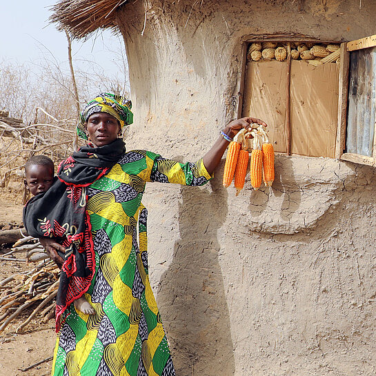 Frau in grün-gelbem Gewand mit einem Kind auf dem Arm und Maiskolben in der Hand vor dem Getreidelager