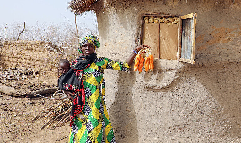 Frau in grün-gelbem Gewand mit einem Kind auf dem Arm und Maiskolben in der Hand vor dem Getreidelager