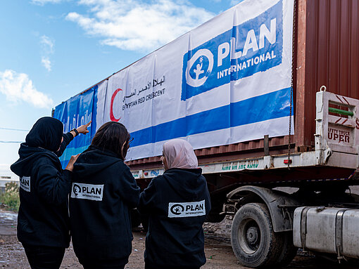 Drei Frauen stehen mit dem Rücken zur Kamera und blicken auf einen LKW, auf dem ein großes Banner mit den Logos von Plan International und dem Internationalen Roten Kreuz zu sehen sind.