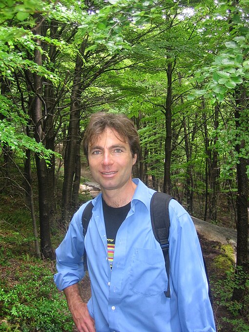 Matthias Knab auf Reisen Mann in blauem Hemd und mit Rucksack steht im Wald