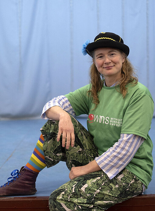 Elin und ihre Kolleginnen bilden auch Interessierte in Clownerie aus. Eine Frau in bunten Anziesachen und einem Hut sitzt auf dem Rand einer Bühne. Sie lächelt freundlich in die Kamera.