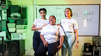 Drei junge Frauen stehen und sitzen gemeinsam in einem Schulungsraum; eine Person sitzt auf einem Hocker, zwei weitere stehen unterstützend daneben, im Hintergrund eine Tafel mit technischen Zeichnungen und Geräteausstattung.