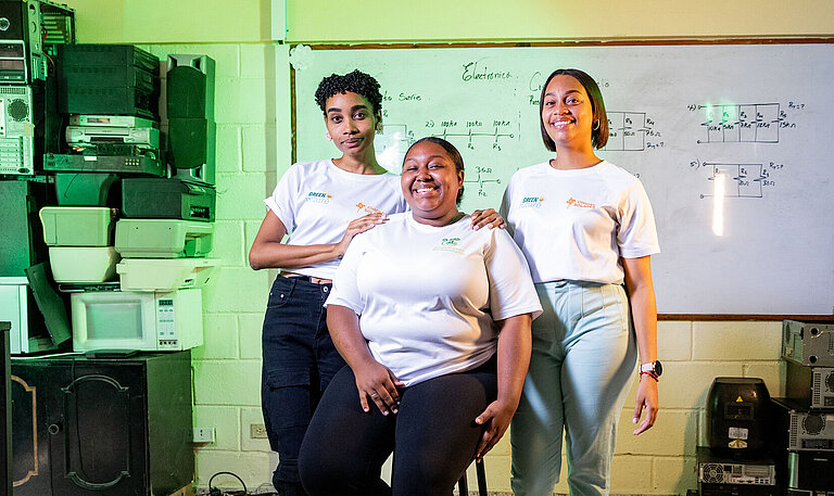 Drei junge Frauen stehen und sitzen gemeinsam in einem Schulungsraum; eine Person sitzt auf einem Hocker, zwei weitere stehen unterstützend daneben, im Hintergrund eine Tafel mit technischen Zeichnungen und Geräteausstattung.