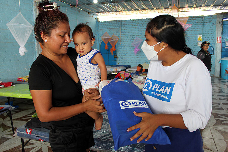 Eine Mitarbeiterin von Plan International reicht einer Mutter mit Baby einen gefüllten Stoffbeutel