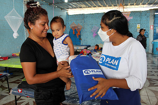 Humanitäre Hilfe Eine Mitarbeiterin von Plan International reicht einer Mutter mit Baby einen gefüllten Stoffbeutel