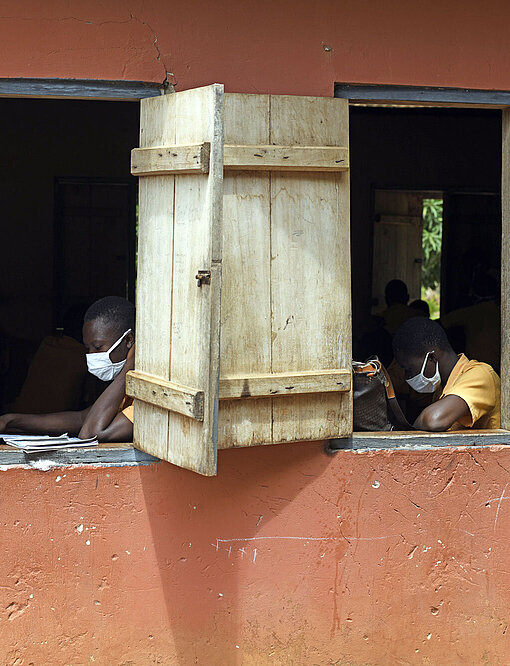 Durch die Corona-Pandemie sind viele Schüler:innen mit dem Lehrplan hinterher. Blick auf zwei offene Fenster eines Klassenzimmers. Darin sitzen Schüler:innen mit Masken und lesen konzentriert.