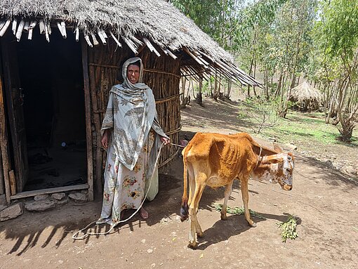 Eine Frau steht vor einer Hütte und hat ein abgemagertes Kalb an einer Leine