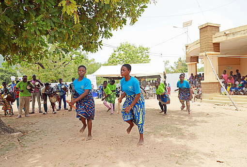 Zwei Mädchen in bunten Kleidern rennen barfuß über einen Sandweg unter Bäumen