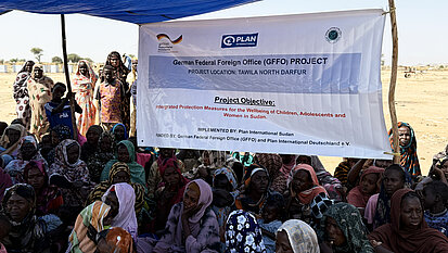 Eine große Gruppe von Frauen und Kindern sitzt unter einem blauen Sonnendach; hinter ihnen hängt ein Banner mit Plan-Logo und Text zu einem Projekt.