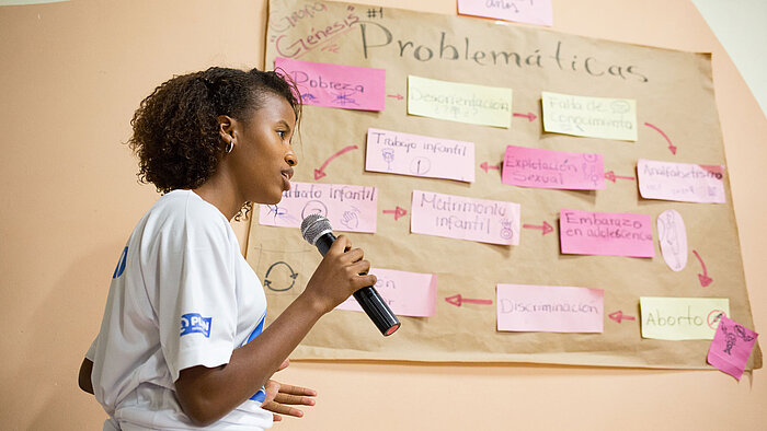 Eine Teenagerin steht neben einem Plakat, was auf Spanisch Probleme in der Gemeinde thematisiert. In ihrer Hand hält sie ein Mikrofon, außerdem trägt sie ein T-Shirt von Plan International