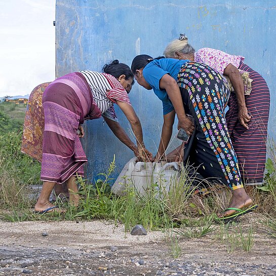 Drei Frauen in bunter Kleidung beugen sich zu einem Brunnen