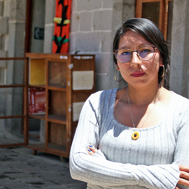 Eine junge Frau in weißem Pullover und mit Brille steht selbstbewusst mit verschränkiten Armen vor einem Gebäude