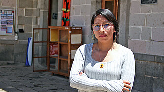 Eine junge Frau in weißem Pullover und mit Brille steht selbstbewusst mit verschränkiten Armen vor einem Gebäude