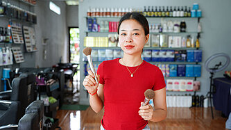 Eine junge Asiatin in rotem T-Shirt steht inmitten eines Friseursalons und schaut in die Kamera