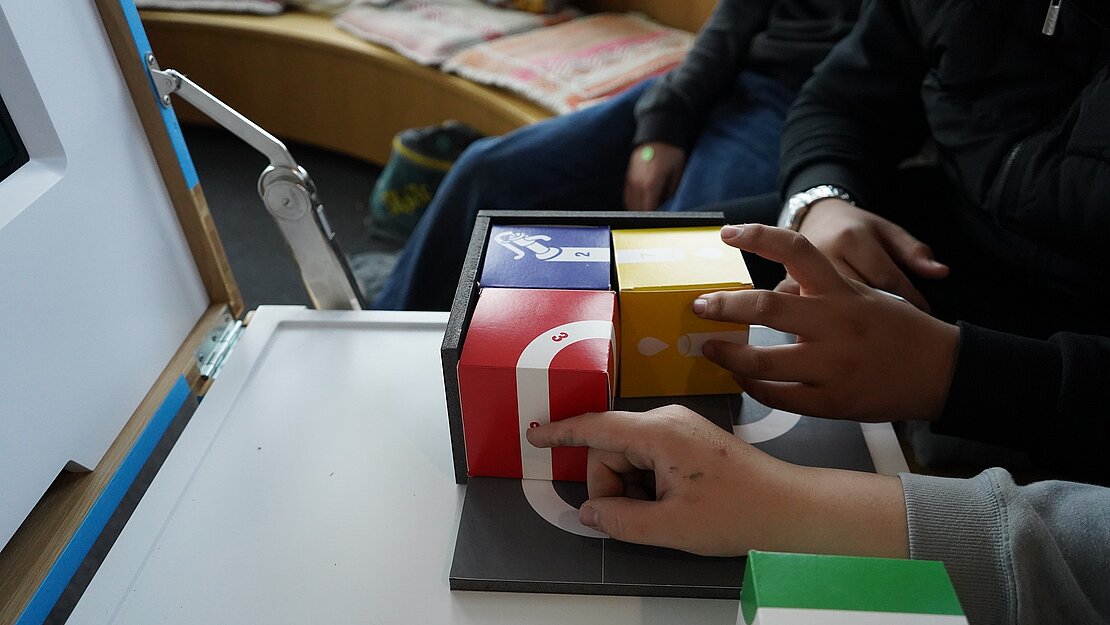 Zwei Jungen, von denen nur die Hände zu sehen sind, legen vier große bunte Würfel in rot, gelb, grün und blau auf einem Spielfeld zusammen. Auf den Würfeln sind weiße Linien eingezeichnet