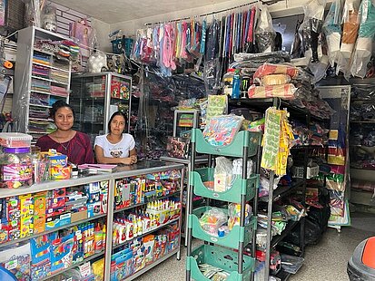 Hinter einer Ladentheke mit zahlreichen bunten Waren stehen zwei junge Frauen und lächeln in die Kamera.