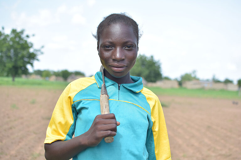 Assétou steht auf einem Feld und hält eine Sichel in der Hand. 