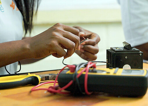 Hände einer Projektteilnehmenden, die Kabel miteinander verbindet; auf dem Tisch liegen ein Messgerät und weitere elektronische Komponenten aus der praktischen Ausbildung.