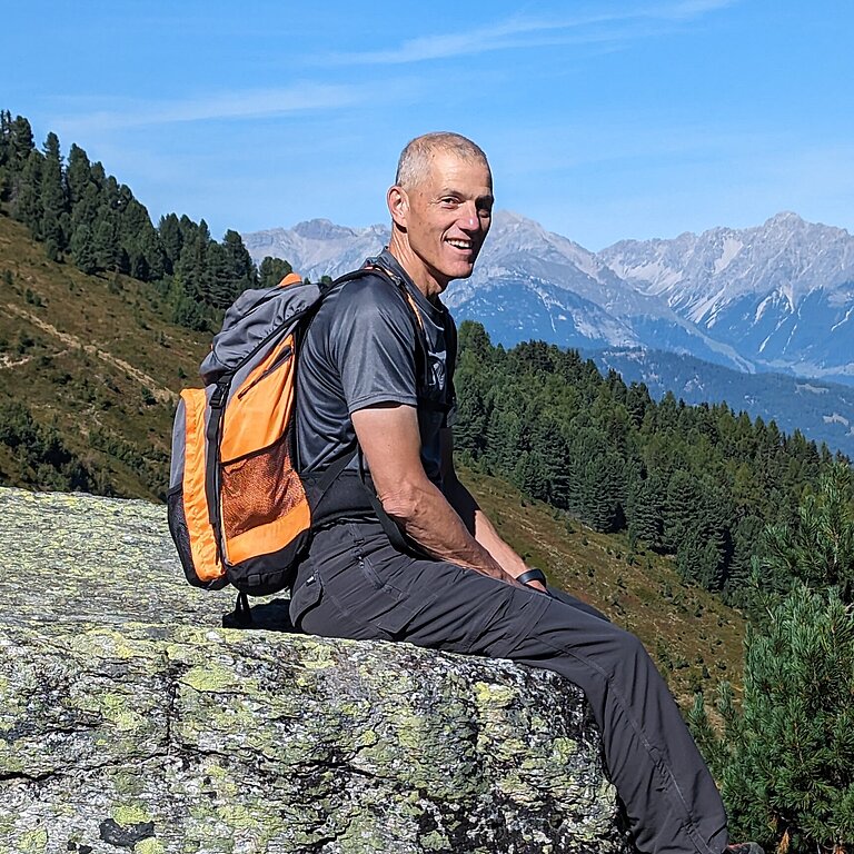 Stifter Herbert Schönebeck sitzt auf einem großen Stein vor eindrucksvoller Bergkulisse
