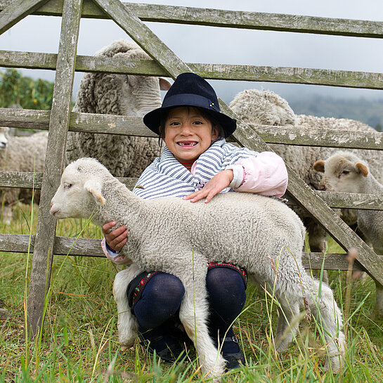 Ein Mädchen sitzt im Gras vor einem Weidezaun und hält ein Lamm im Arm