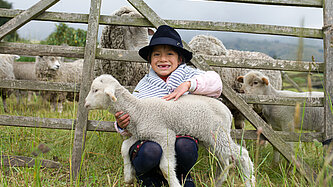 Ein Mädchen sitzt im Gras vor einem Weidezaun und hält ein Lamm im Arm