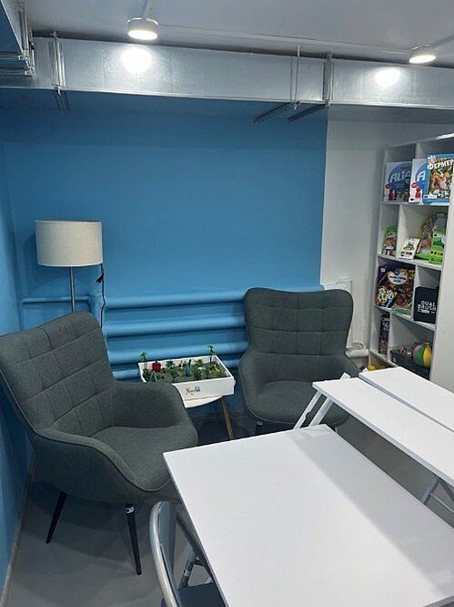 Ein kleines Zimmer ohne Fenster mit einem Tisch, zwei Sesseln, einer Lampe und einem Regal