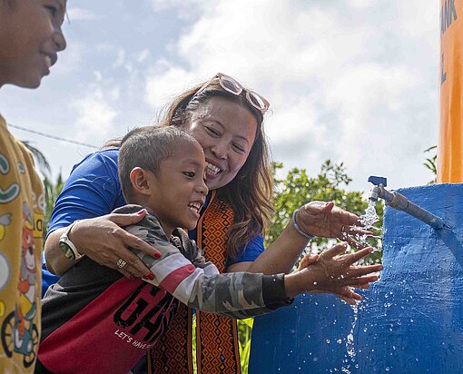 Wasser marsch: Endlich kommt die wertvolle Ressource direkt in die Dörfer Eine Frau steht mit Kindern an einem Wasserhahn und lacht