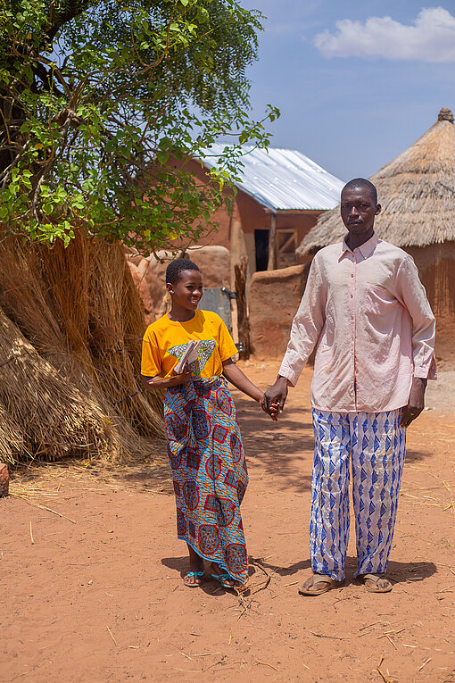 Ein ghanaisches Mädchen im Teenageralter und ihr Vater stehen vor einem Baum und Lehmhütten und halten sich an der Hand