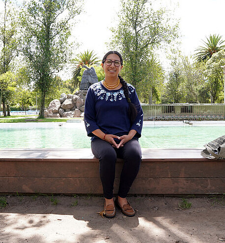 Verónica steht im Freien vor einer Wasserquelle in einer ländlichen Umgebung und blickt in die Kamera