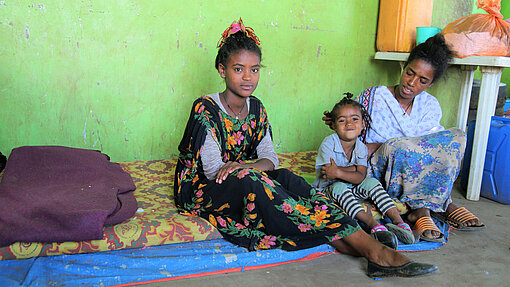 Eine Mutter sitzt mit zwei Kindern auf einer Matratze vor einer grünen Wand. Alle drei blicken in die Kamera; das jüngste Kind lächelt. Neben ihnen stehen Wasserkanister und Haushaltsgegenstände.