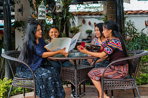 Vier junge Frauen sitzen um einen runden Tisch herum. Eine von ihnen liest ein Dokument, die anderen sprechen miteinander