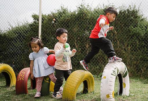 Drei Kinder spielen mit bunten Bällen und laufen über bemalte Reifen im Freien. Ein Kind balanciert auf einem Reifen, während die anderen zuschauen.