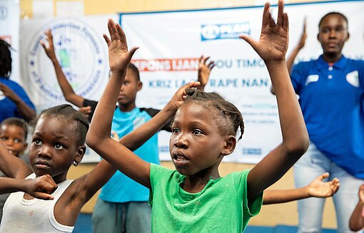 Plan International hat in haitianischen Lagern für geflüchtete Menschen sichere Räume eingerichtet, in denen Kinder geschützt lernen, spielen und ihre freie Zeit verbringen können Ein Mädchen tanzt mit anderen Kindern