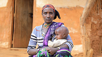 Eine Frau sitzt vor einer Lehmhütte, sie hält ein Kleinkind im Arm und schaut ernst in die Kamera