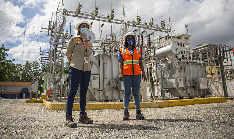 Zwei Frauen in Arbeits-Sicherheitskleidung stehen vor einem Kraftwerk
