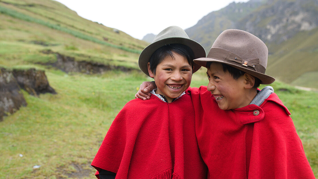 Zwei Jungen in roten Ponchos stehen auf einer Wiese im Hochland, haben den Arm umeinander gelegt und lachen