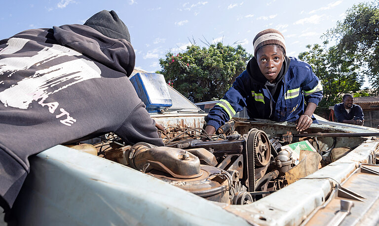 Zwei junge Frauen in Overall beugen sich über den Motorblock eines alten Autos
