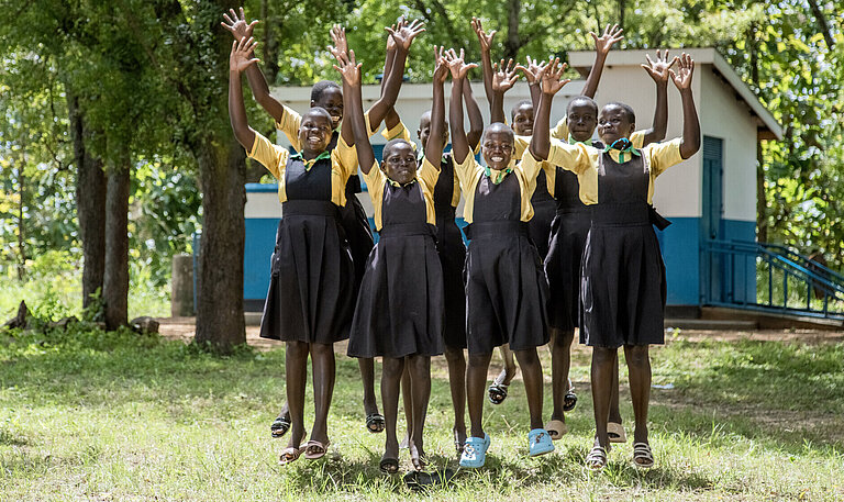 Gruppe von fröhlichen Schülerinnen in Schuluniformen springt mit erhobenen Armen auf einer Wiese vor einem Schulgebäude in Uganda.