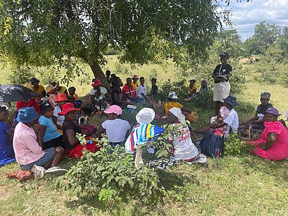 Eine Gruppe von Frauen sitzt im Kreis unter einem Baum auf dem Boden. Die Frauen tragen verschiedene Hüte und bunte Kleidung. Eine Person steht in der Mitte und hält etwas in der Hand, während sie spricht oder Anweisungen gibt.