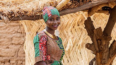 Eine junge Frau aus Nigeria steht draußen, sie trägt ein buntes Kleid und ein Tuch um das Haar gewickelt und lächelt in die Kamera