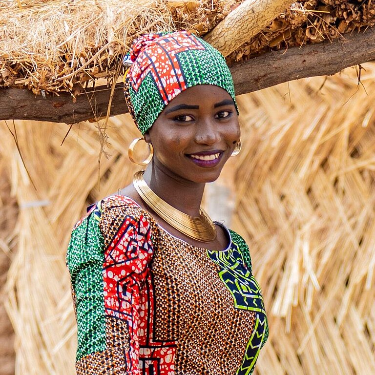 Eine junge Frau aus Nigeria steht draußen, sie trägt ein buntes Kleid und ein Tuch um das Haar gewickelt und lächelt in die Kamera
