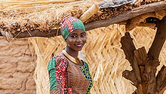 Eine junge Frau aus Nigeria steht draußen, sie trägt ein buntes Kleid und ein Tuch um das Haar gewickelt und lächelt in die Kamera