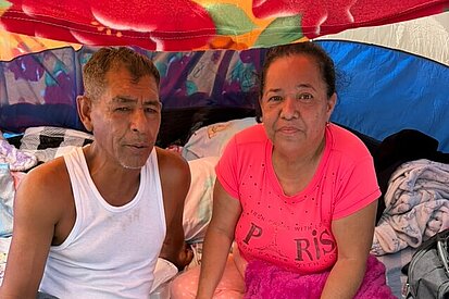 Ein älteres Paar sitzt in einem Zelt vor ihrer beschädigten Wohnung in Caracas und blickt in die Kamera; ihr Hab und Gut ist um sie herum verteilt.