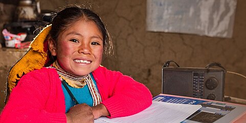 Ein Mädchen sitzt an einem Schreibtisch. Vor ihr liegt ein aufgeklapptes Heft, ein Ordner und ein Radio. Sie grinst in die Kamera und hält einen Stift in der Hand. 