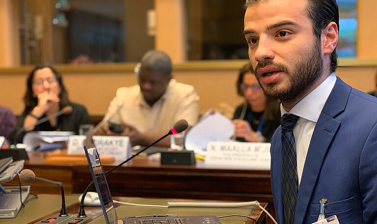 Karim sitzt in einer Konferenz und spricht in ein Mikrofon.