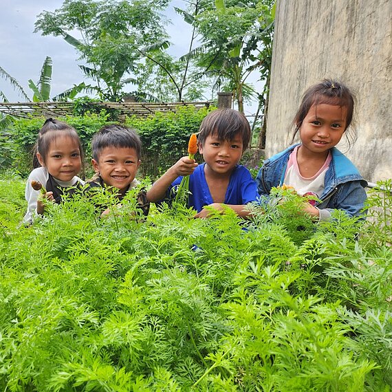 Vier Kinder sitzen lächelnd in einem üppigen grünen Gemüsegarten. Ein Junge zeigt eine frisch geerntete Karotte, die anderen Kinder halten ebenfalls Gemüse in den Händen. Im Hintergrund sind Pflanzen, Bäume und ein Teil einer Mauer zu sehen.