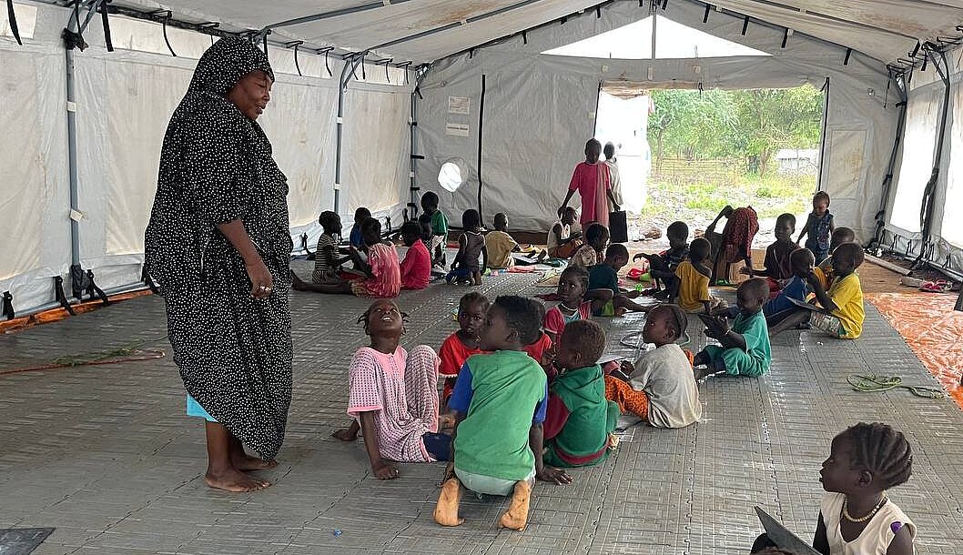 Kinder mit Lehrerin im Geflüchtetencamp Viele Kinder sitzen in einem großen Zelt auf dem Boden, eine Frau steht in der Mitte und spricht mit einer Gruppe Kinder