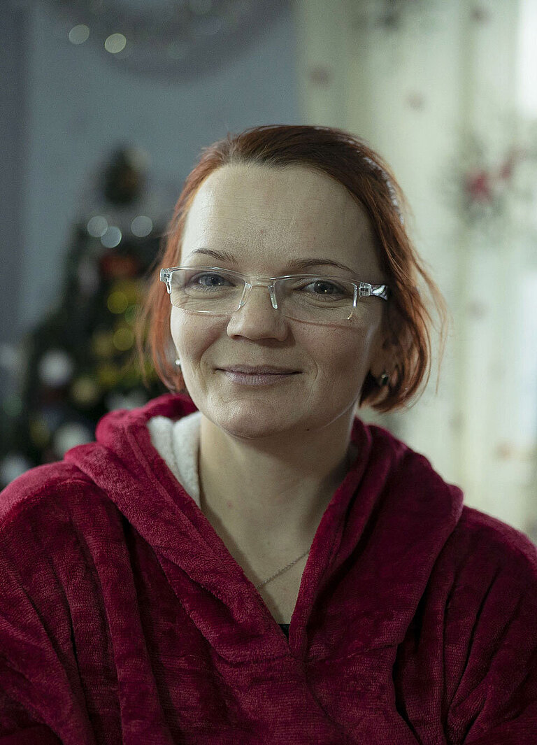 Eine Frau mit Brille und rotem Kapuzenpullover schaut in die Kamera. Sie befindet sich in einem Zimmer.