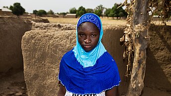 Ein afrikanisches Mädchen mit leuchtend blauem Kopftuch steht vor einer halbhohen Mauer aus Lehm und Stein, im Hintergrund sieht man einige Hütten aus ihrem Dorf.