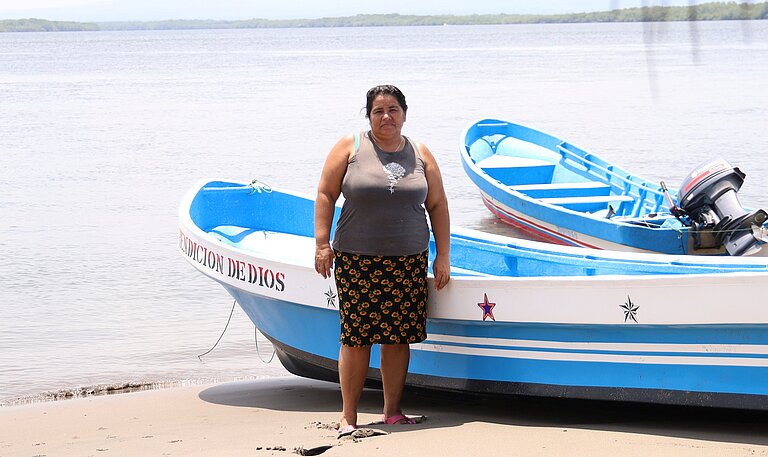 Eine Frau steht am Strand, im Hintergrund ist ein Fischerboot am Ufer zu sehen.